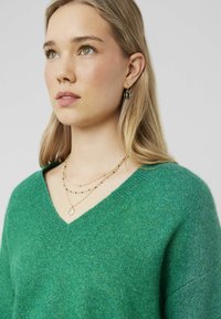 Pull vert à col en V en maille douce, accompagné de colliers en or superposés avec des perles noires et un charm, ainsi que de petites boucles d'oreilles au design circulaire.