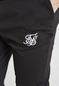 Schwarze Sporthose mit glatter Textur, die ein weiß gesticktes Logo am linken Oberschenkel aufweist. Die Hose hat seitliche Taschen zur Aufbewahrung.