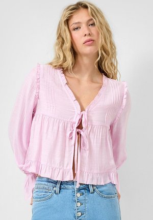 Blonde Frau mit welligem Haar, die eine hellrosa langärmelige Bluse mit Frontbändern und Rüschendetails trägt, kombiniert mit hoch taillierten blauen Jeans.