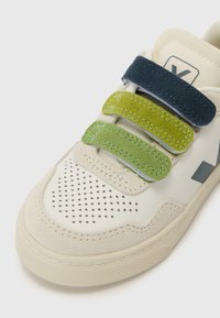 Nærbilde av kremfarget sneakersko med perforert tå, utstyrt med tre borrelåser i mørkeblått, grønt og lysegrønt, samt teksturerte detaljer.