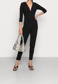 Woman wearing a black V-neck jumpsuit and black heeled sandals, holding a white and black snakeskin-patterned handbag.