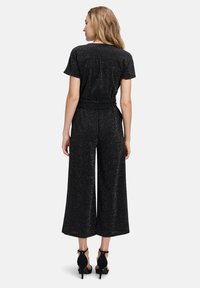 Combinaison noire en tissu texturé et étincelant, avec des manches courtes, une taille cintrée, un pantalon évasé et une silhouette décontractée.