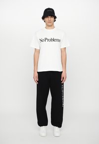 White cotton t-shirt with "No Problemo" in black text, paired with black sweatpants featuring "No Problemo" printed vertically, worn with white shoes.