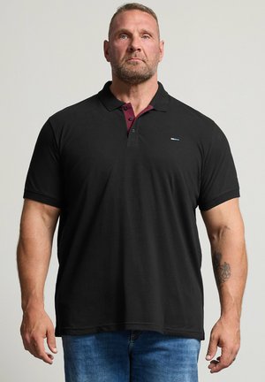 Muskelmand med kort hår og skæg iført sort poloshirt med bordeaux detaljer på kraven og blå jeans, stående mod en ensfarvet baggrund.