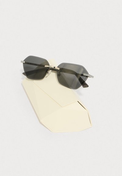 Lunettes de soleil angulaires gris foncé avec des accents argentés reposant sur un étui géométrique beige, présentant une texture lisse et un design minimaliste.