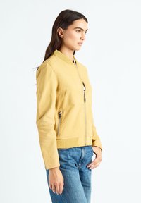 Blouson aviateur en cuir jaune avec fermeture éclair à l'avant, col côtelé et poches latérales; porté avec un jean bleu sur un fond neutre.