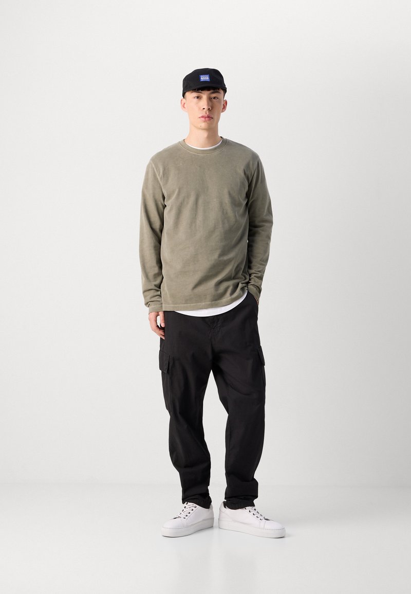 Chemise à manches longues vert olive, pantalon cargo noir, baskets blanches, casquette noire avec un patch logo bleu, se tenant contre un fond neutre.