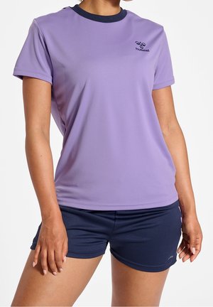 Kvinde iført en kortærmet lavendelfarvet sportstrøje og marineblå atletikshorts, stående med den ene hånd afslappet ned langs siden.