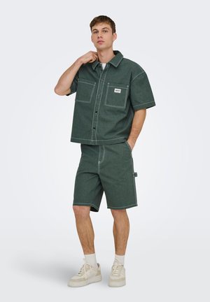 Tânăr purtând cămașă verde închis cu mâneci scurte și nasturi și pantaloni scurți asortați, stând cu o mână în buzunar și adidași albi.