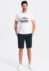 T-shirt en coton blanc avec "OMBRE UNIVERSITY" imprimé en noir, associé à un short bleu marine et des baskets blanches. Manches courtes, coupe décontractée.