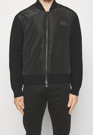 Czarna kurtka bomberka z błyszczącym nylonowym frontem, rękawami i kołnierzem z prążkowanego dzianiny, wyposażona w centralny zamek błyskawiczny i boczne kieszenie. Subtelne detale z logo.