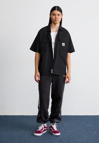 Svart kortermet skjorte med knapper, to frontlommer og logo-patch, sammen med grå joggesbukser med en subtil sideprint og røde joggesko.