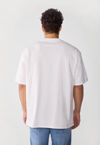 Hvit bomulls t-skjorte med korte ermer, avslappet passform og synlige sømdetaljer langs ermene og nederkant. Brukt sammen med blå denimjeans.