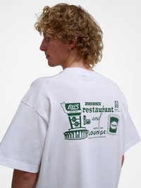 Ung person med krøllete blondt hår iført en hvit oversized t-skjorte med grønn grafisk tekst og bilder på ryggen.