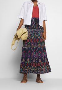 Maxikjol i marinblått med flerfärgade geometriska mönster, lagerdesign, tillsammans med en vit skjorta och en ljusgul väska. Bruna sandaler.