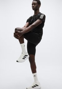 Μαύρο αθλητικό μπλουζάκι με τρεις λευκές ρίγες, μαύρο σορτς, λευκά αθλητικά παπούτσια με μαύρα στοιχεία και λευκές κάλτσες, σε στάση διατάσεων άσκησης.