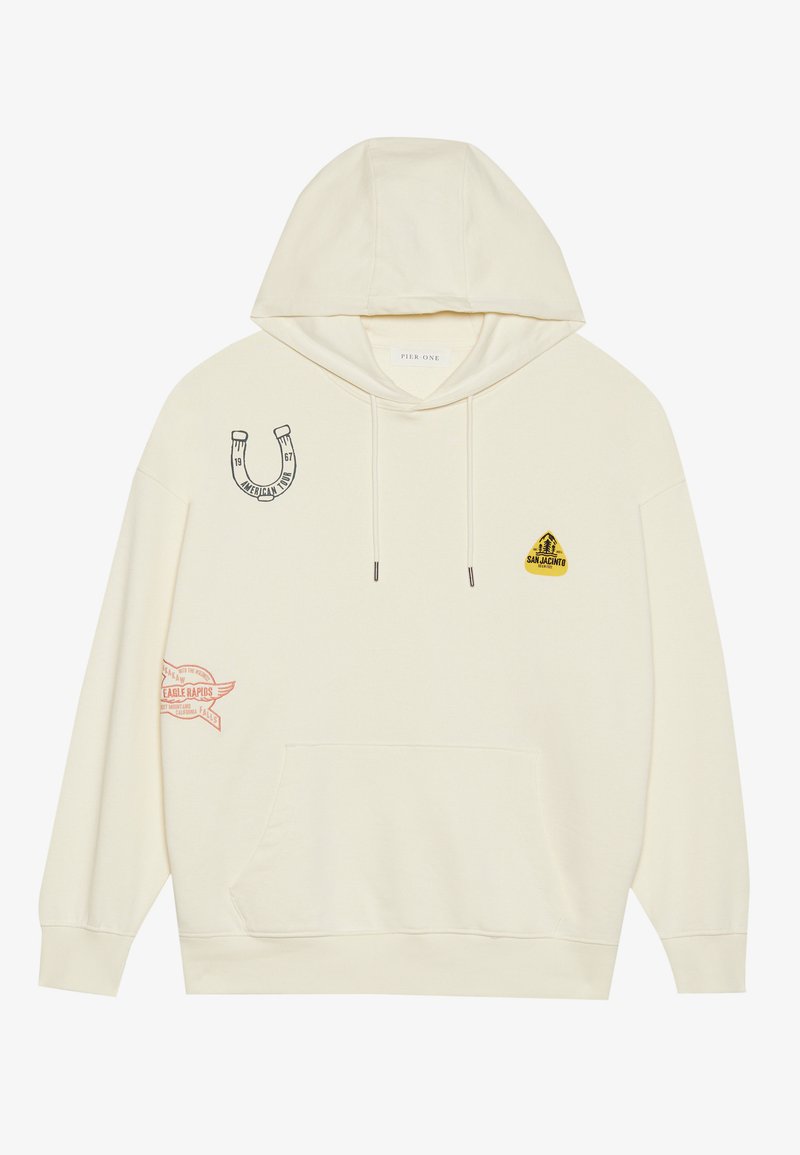 Haut à capuche couleur crème avec une poche avant, orné de divers écussons : un fer à cheval, un triangle jaune et une bannière rouge avec du texte. En coton.