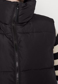 Czarna kamizelka puchowa z wysokim kołnierzem, zapięciem na zamek z przodu i pikowanym przeszyciem. Materiał wydaje się gładki i ocieplony.