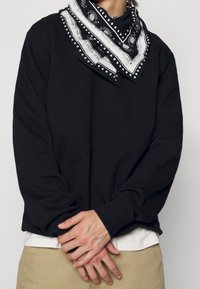 Personne portant une chemise noire à manches longues, un pantalon beige et un bandana à motif noir et blanc autour du cou, mains jointes devant.