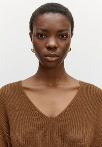 Brauner Strickoberteil mit V-Ausschnitt, das ein strukturiertes Muster und sichtbare Nähte aufweist. Das Modell trägt goldene kreisförmige Ohrringe.