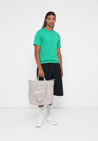 Γκρι νάιλον τσάντα ώμου με λευκό λογότυπο Nike, συνδυασμένη με πράσινο μπλουζάκι και μαύρη βερμούδα. Το μοντέλο στέκεται μπροστά σε λευκό απλό φόντο.