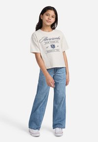 T-shirt cropped a maniche corte di un beige chiaro con stampa grafica blu navy. Abbinato a jeans di denim blu a gamba larga e sneakers bianche.