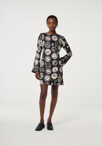 Schwarzes Kleid mit weißen kreisförmigen Mustern, langen Glockenärmel und geradem Schnitt. Getragen mit schwarzen Slippern, glatte Stofftextur.