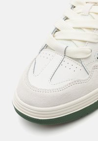 Hvide lædersneakers med grå ruskind og grøn gummisål, der har perforerede detaljer og lys beige snørebånd.