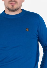 Mężczyzna w niebieskim swetrze z długim rękawem, z małą czarną i złotą naszywką na lewej piersi, na jednolitym białym tle.