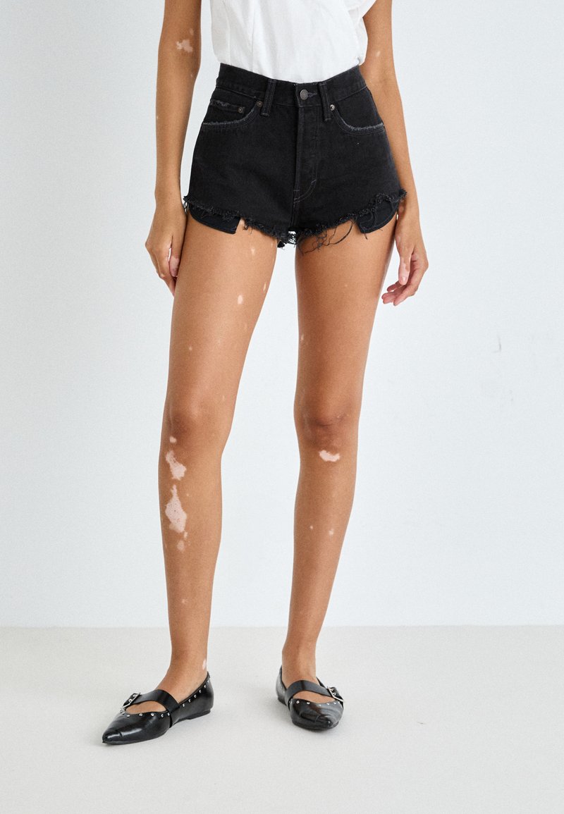 Shorts en denim noir taille haute avec ourlet effiloché, dotés de cinq poches et de matériel en métal, associés à des chaussures plates noires et un top blanc.