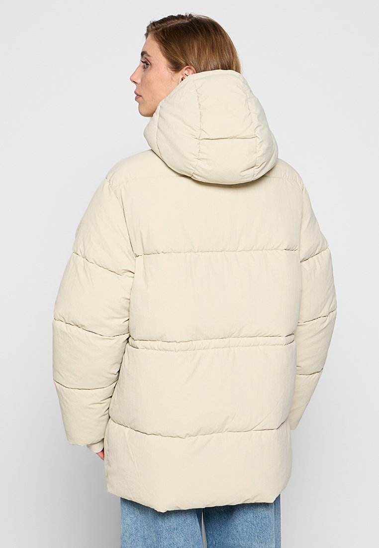 Helle beige Puffy-Jacke mit Kapuze, mit horizontalen, gesteppten Abschnitten und einer lockeren Passform. Glatte Textur, baumwollähnliches Material.
