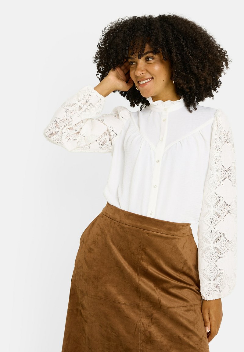 Blouse blanche avec des manches ballons en dentelle, fermeture à boutons et col montant, associée à une jupe en daim marron, au texture lisse et à la coupe ajustée.