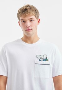 Witte katoenen t-shirt met een zak en een geborduurd olifantenontwerp in blauw en groen. Eenvoudige ronde hals, korte mouwen.