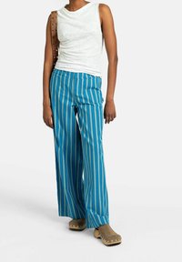 Person med ärmlös vit topp, blå och svarta vertikala randiga vida byxor, brun läderaxelväska och beigefärgade träskor med metallspik.