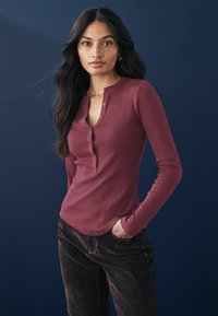 Langærmet, ribbet henley skjorte i burgunder med knaplukning ved halsen, parret med mørkebrune fløjl bukser. Struktureret stof, tætsiddende design.