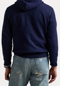 Námořnický modrý mikina s měkkou texturou a přizpůsobeným pasem. Nosená s roztrhanými modrými džíny se viditelným šitím a kapsami.