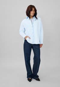 Light blue button-up shirt with long sleeves, paired with dark blue straight-leg jeans and pointed black shoes. Simple design and relaxed fit.