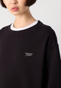 Juodas džemperis su baltu gumele apipintu kaklu ir mažuoju balto siuvinėto "tommy jeans" logotipu krūtinėje. Minkšta audinio tekstūra.