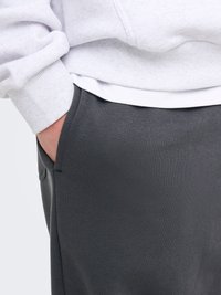 Oseba v temnosivih hlačah z desno roko v žepu, svetlo sivi jopa in belo srajco, vidno na spodnjem robu.