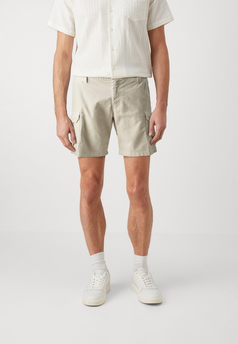 Pantalones cortos cargo de color beige claro, hechos de algodón, con dos bolsillos laterales, corte recto y cierre de botón. Se usan con zapatillas blancas y calcetines.