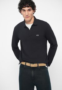 Jovem com cabelo encaracolado vestindo uma polo preta de manga longa com o logo A|X, jeans escuros e um cinto bege, posando contra um fundo liso.