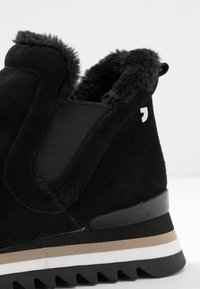 Bottine en daim noir avec doublure en fausse fourrure, panneaux élastiques sur les côtés, et une semelle en caoutchouc épaisse à rainures avec des accents beige et blanc.