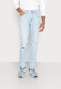 Jasnoniebieskie dżinsy o slim fit z wyrwanym detalem na kolanie. W połączeniu z szarymi butami sportowymi o teksturowanym wzorze.