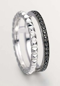 Dos anillos: uno plateado, con un diseño engastado, y otro plateado con una fila de gemas negras montadas en una banda lisa. Ambos presentan acabados pulidos.