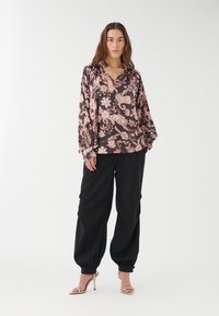 Blusa a maniche lunghe con motivo floreale in nero, rosa e crema; abbinata a pantaloni neri larghi e sandali con tacco nudo.