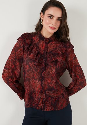 Red paisley-patterned blouse featuring sheer fabric, ruffled collar, and button-down front. Long sleeves with buttoned cuffs.
