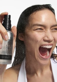 Hydro-Surge Dewy Face Mist in einer klaren Flasche mit schwarzem Pumpkopf, der einen feinen Nebel ausstößt. Der Hintergrund ist in einer hellen, hellen Farbe gehalten und lenkt den Fokus auf das Spray.