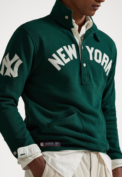 Muž v tmavozelenom svetri s nápisom "NEW YORK" a logom na rukáve, v bielej košeli a svetlých nohaviciach, s rukami v vreckách.