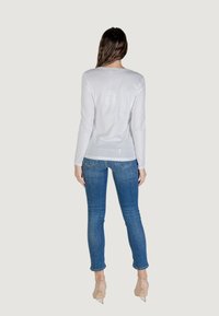 Donna vista da dietro, che indossa un top bianco a maniche lunghe e jeans blu, in piedi su uno sfondo neutro.