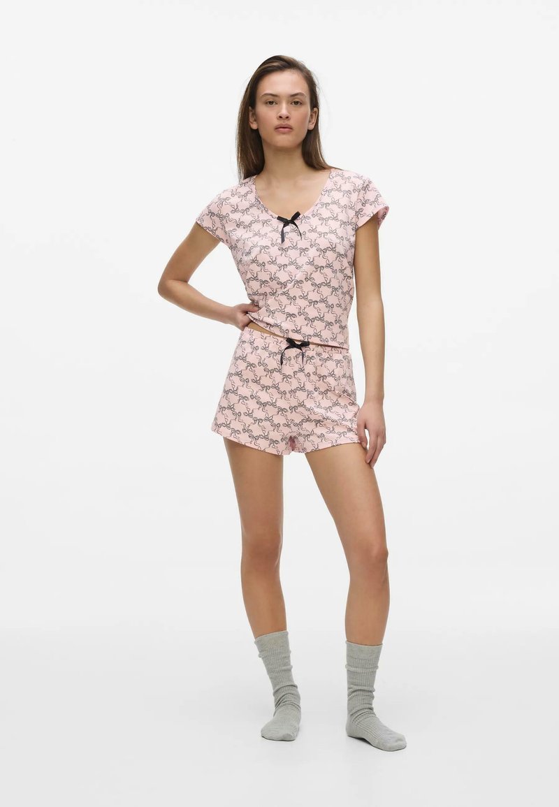 Jeune femme debout, main sur la taille, portant un haut à manches courtes rose à motifs et un short assorti avec des chaussettes grises sur fond blanc.